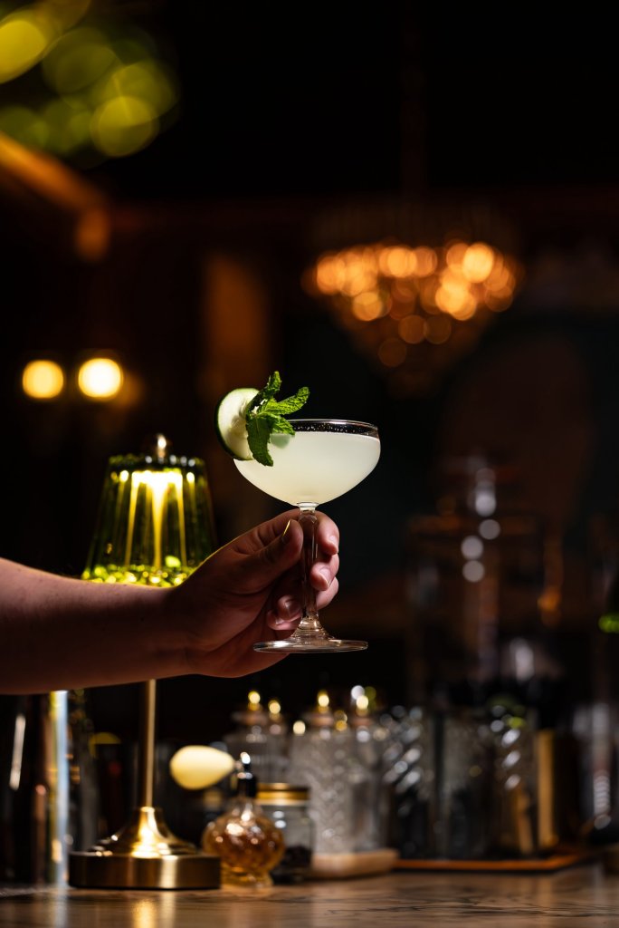 Pale green cocktail in a coupe garnished with cucumber and mint, held up at the bar