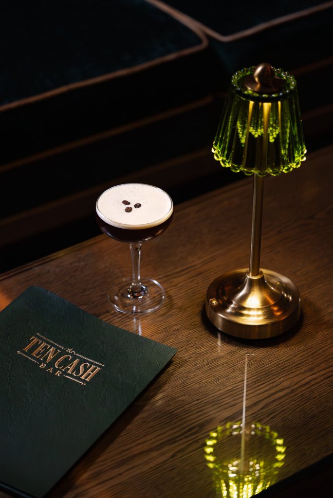 Espresso martini in a coupe topped with three coffee beans, beside a green glass lamp and the Ten Cash menu