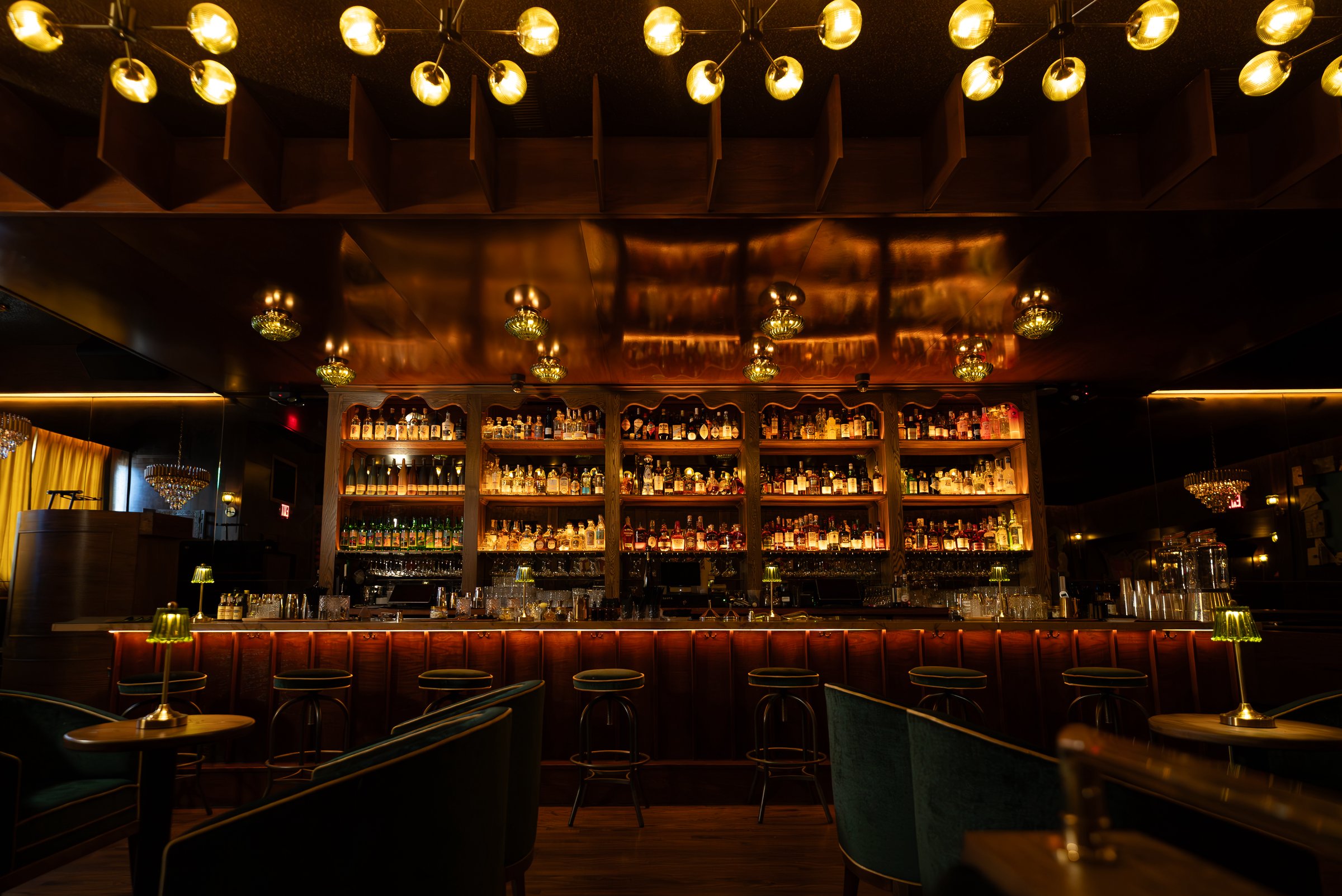 The back bar at Ten Cash Bar, lit in warm gold under brass chandeliers, with green velvet seating