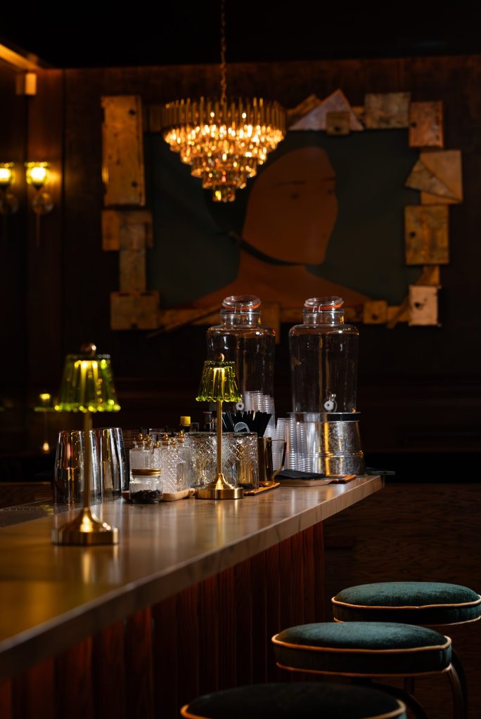 Bar rail with glassware, a green glass lamp and portrait art behind the bar